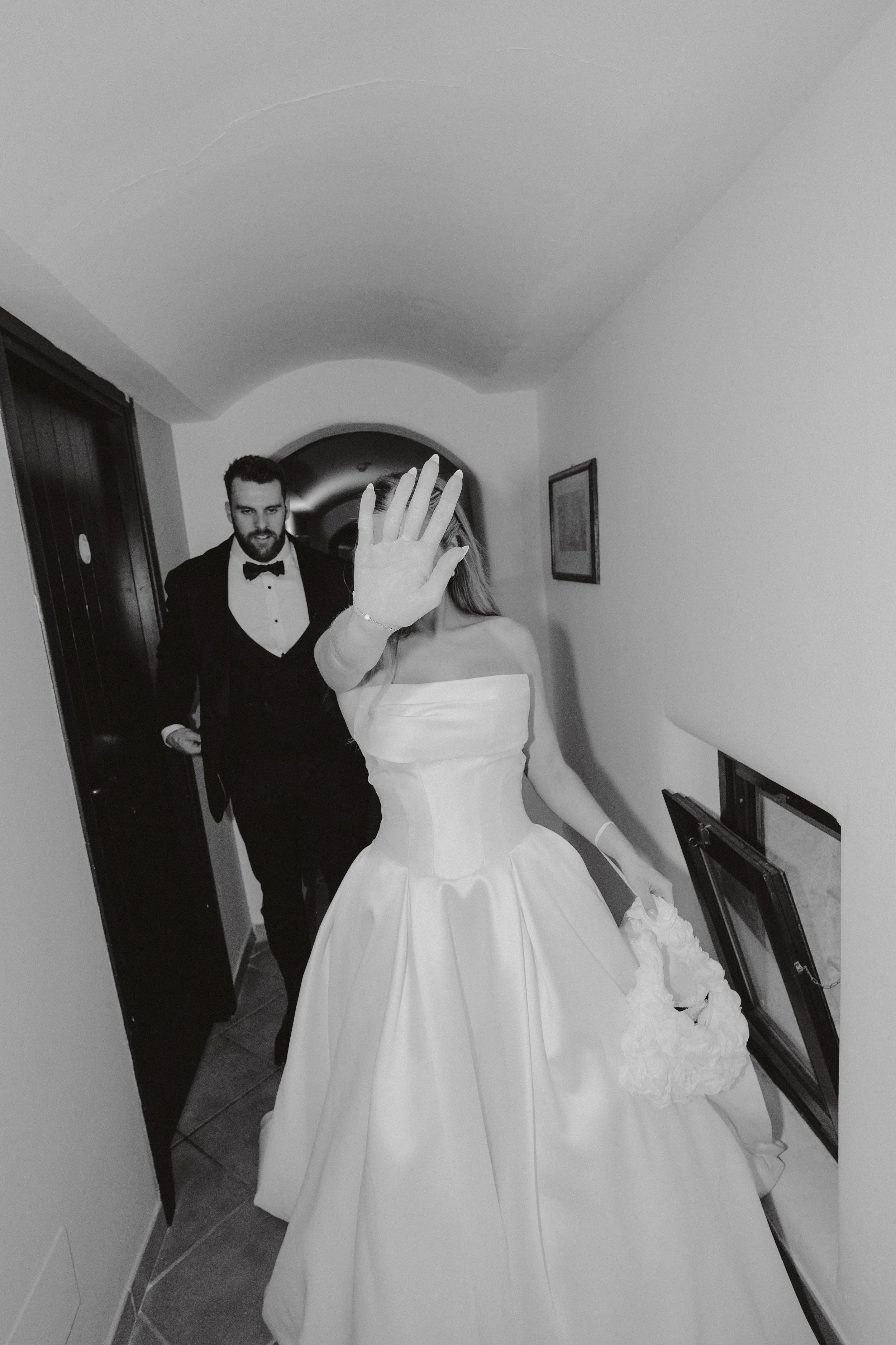 A bride in a strapless wedding dress holding a bouquet, with her hand raised in front of her face, and a groom dressed in a tuxedo walking behind her in a hallway.