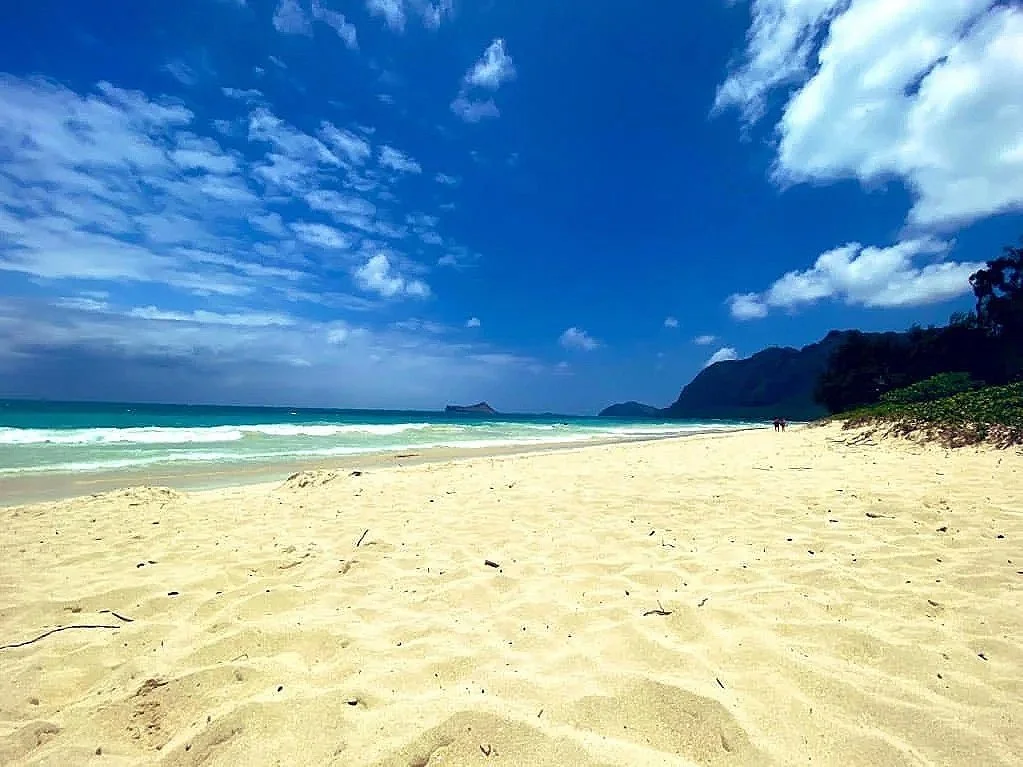 Empty sandy beach with waves crashing on the shore, a blue sky with scattered clouds, and distant mountains covered with greenery.