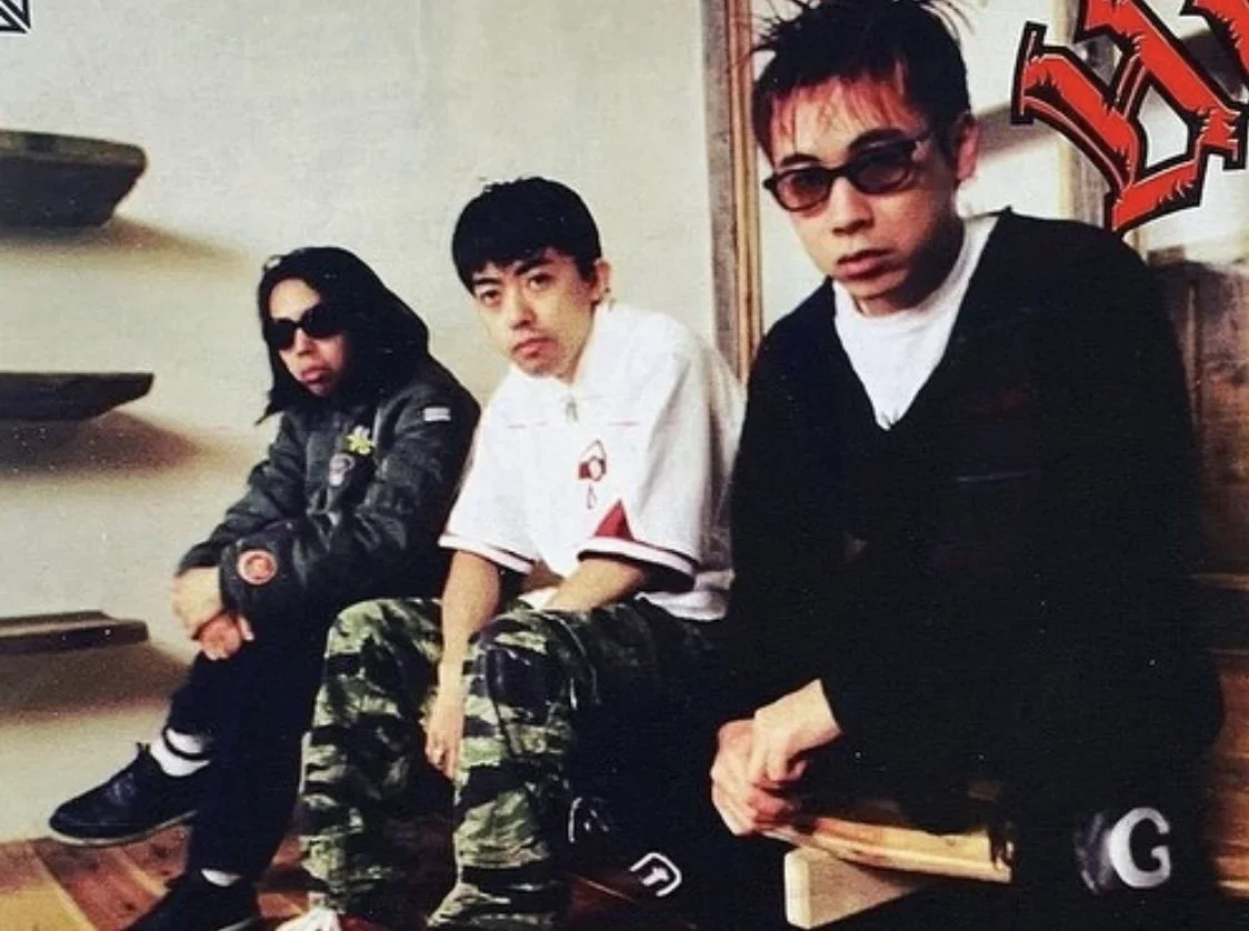 An archival 1990s photo of the Ura-Harajuku pioneers: Jun Takahashi (left), NIGO (center), and Hiroshi Fujiwara (right) sitting together.