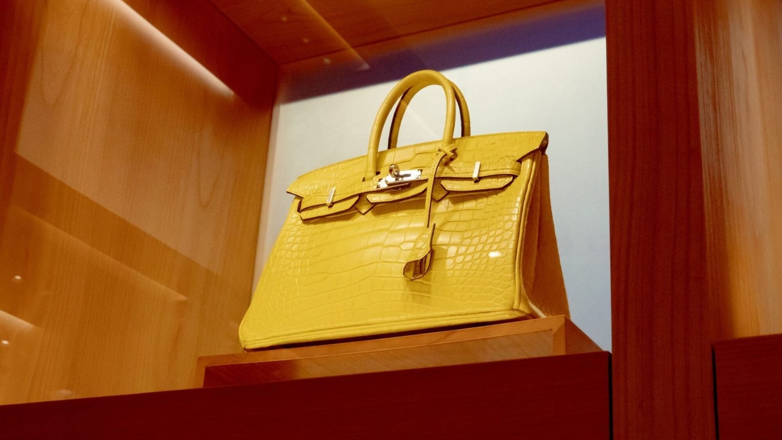 A yellow crocodile Hermes Birkin bag displayed on a wooden shelf under light, highlighting its status as an object for viewing rather than for use.