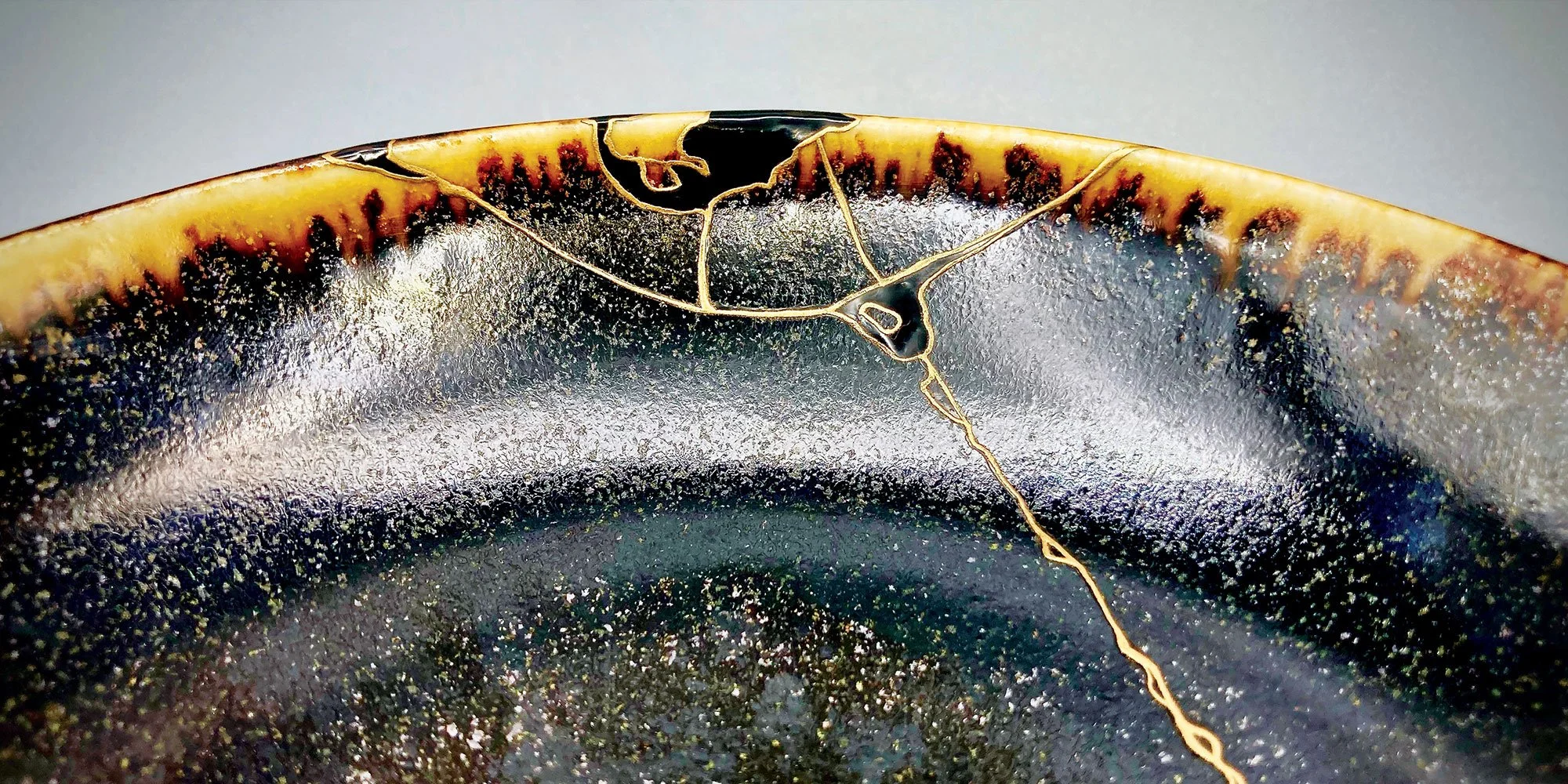 A close-up photograph of a dark ceramic bowl repaired with kintsugi. Delicate lines of gold lacquer trace the cracks, highlighting the breakage as a beautiful feature against the bowl's speckled, glazed surface.