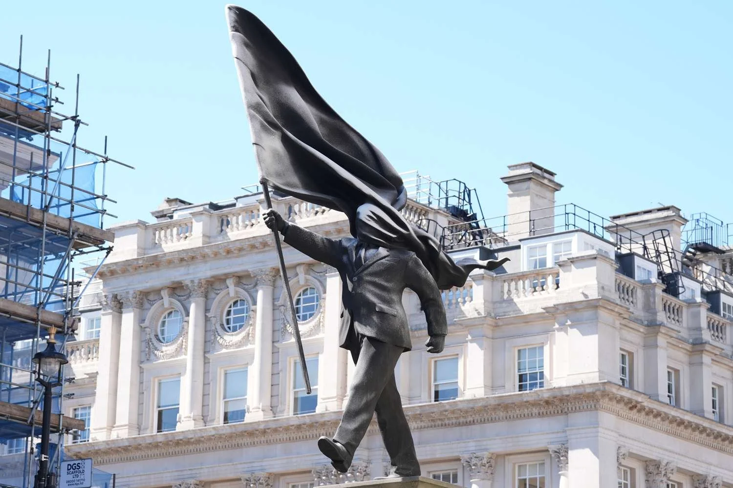A 25-foot bronze and granite statue of a suited man walking off a stone plinth in London's Waterloo Place, his face entirely obscured by a large billowing flag.