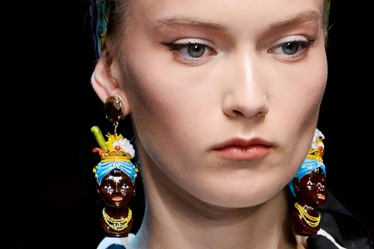 A close-up of a model wearing Dolce & Gabbana earrings featuring "Blackamoor" figures, a controversial accessory from the 2013 collection rooted in colonial-era servitude.