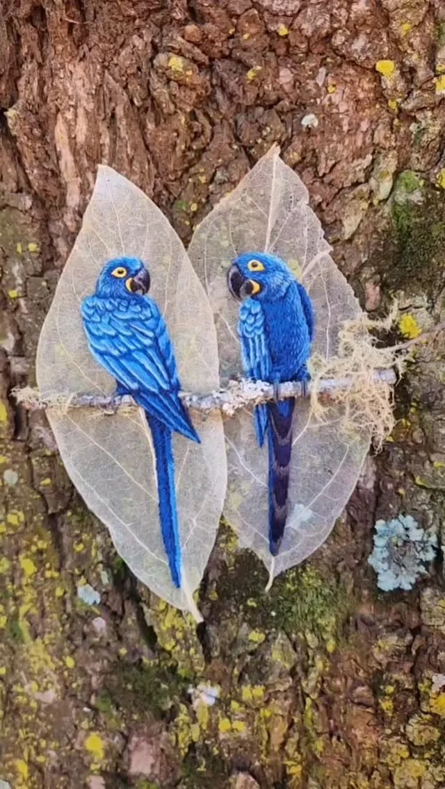 Two vibrant cobalt blue Hyacinth Macaws (Arara-azul) embroidered on skeletonized leaves. The Chromatic Weight of the color is striking against the natural texture of the tree bark, symbolizing environmental resilience in the Pantanal.