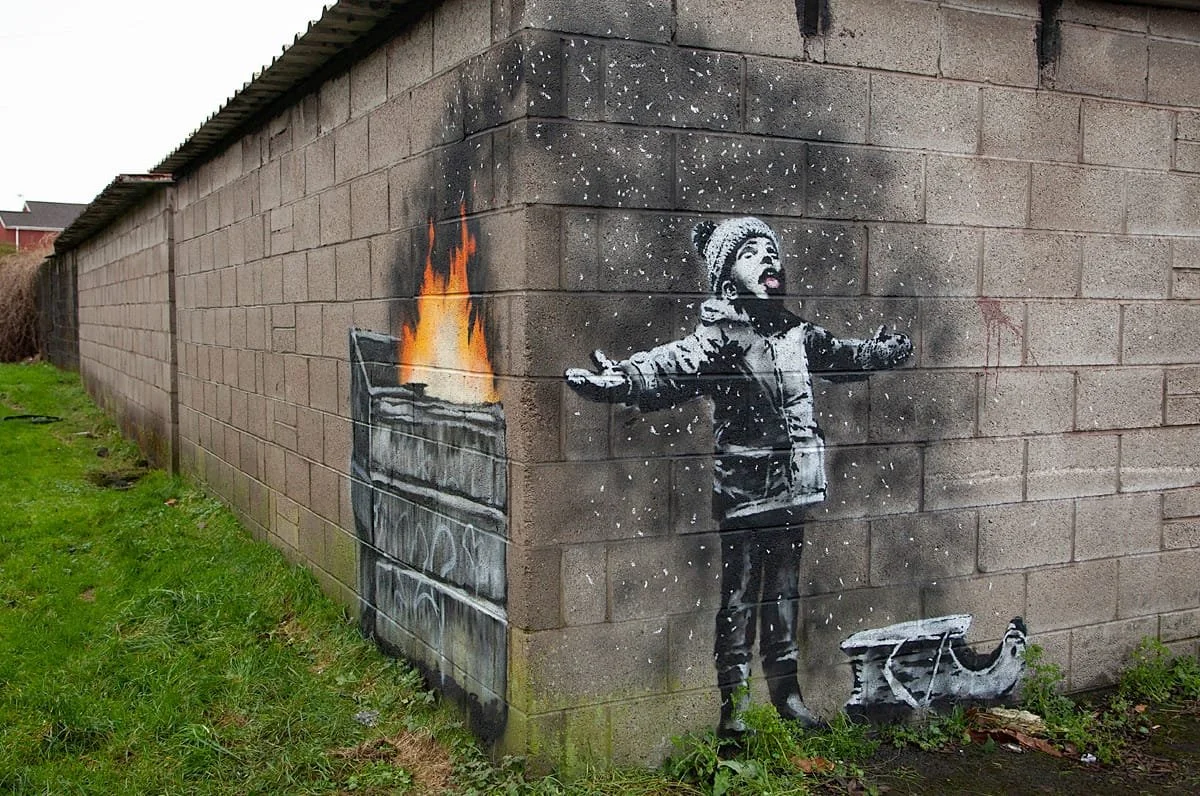 Banksy's 'Season's Greetings' mural on a cinder block wall in Port Talbot, depicting a child tasting falling ash from a burning dumpster, illustrating the PLCFA framework's 'Liquidation of the Simulacrum.