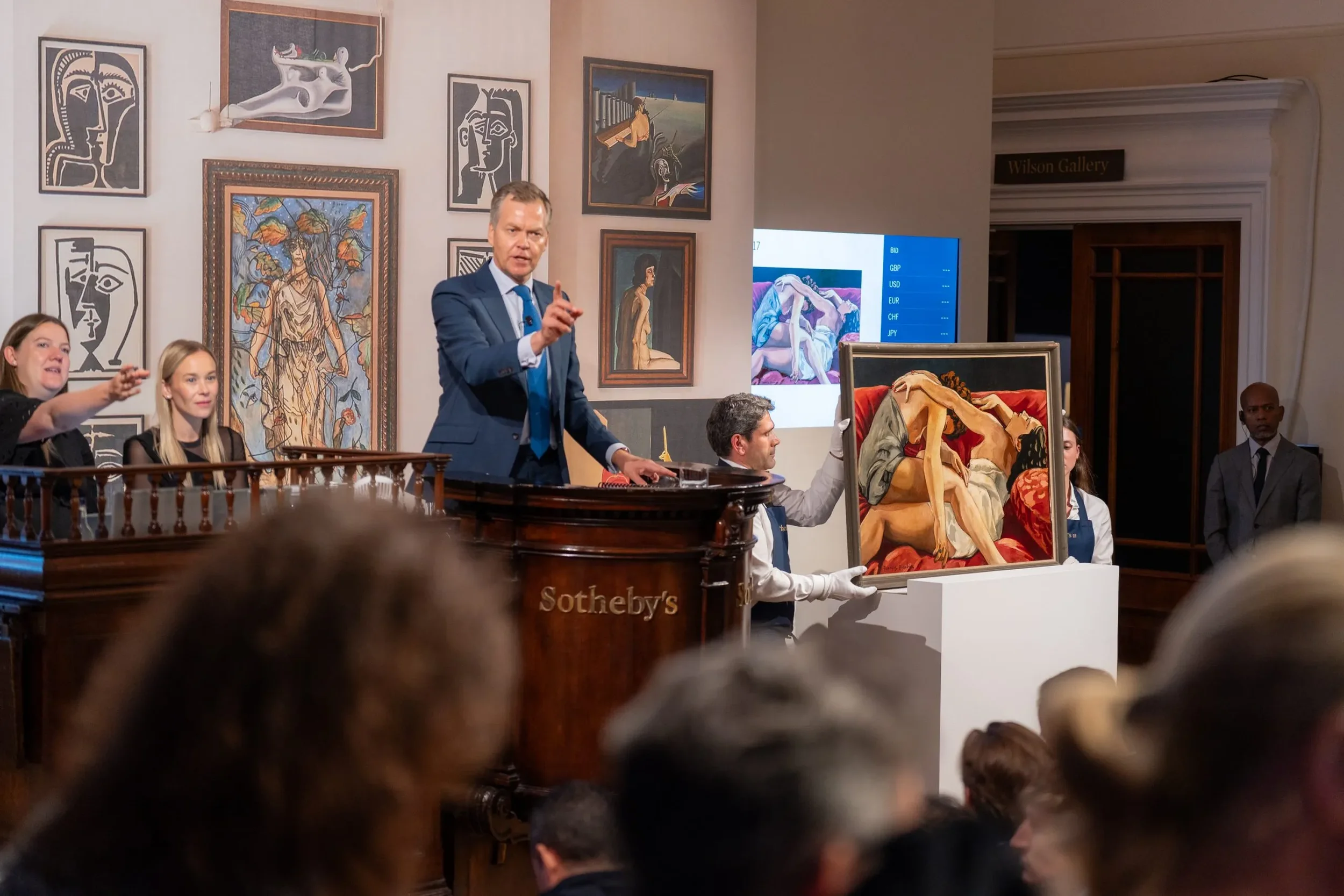 An auctioneer in a blue suit gestures from a wooden Sotheby's podium during an evening sale, while a white-gloved art handler presents a figurative painting of two intertwined figures on a white plinth.