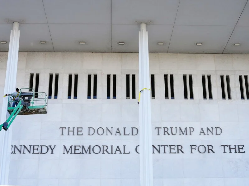 A construction worker in a green scissor lift installs large bronze lettering on the white marble facade of the Kennedy Center, modifying the name to read "THE DONALD J. TRUMP AND JOHN F. KENNEDY MEMORIAL CENTER FOR THE PERFORMING ARTS."
