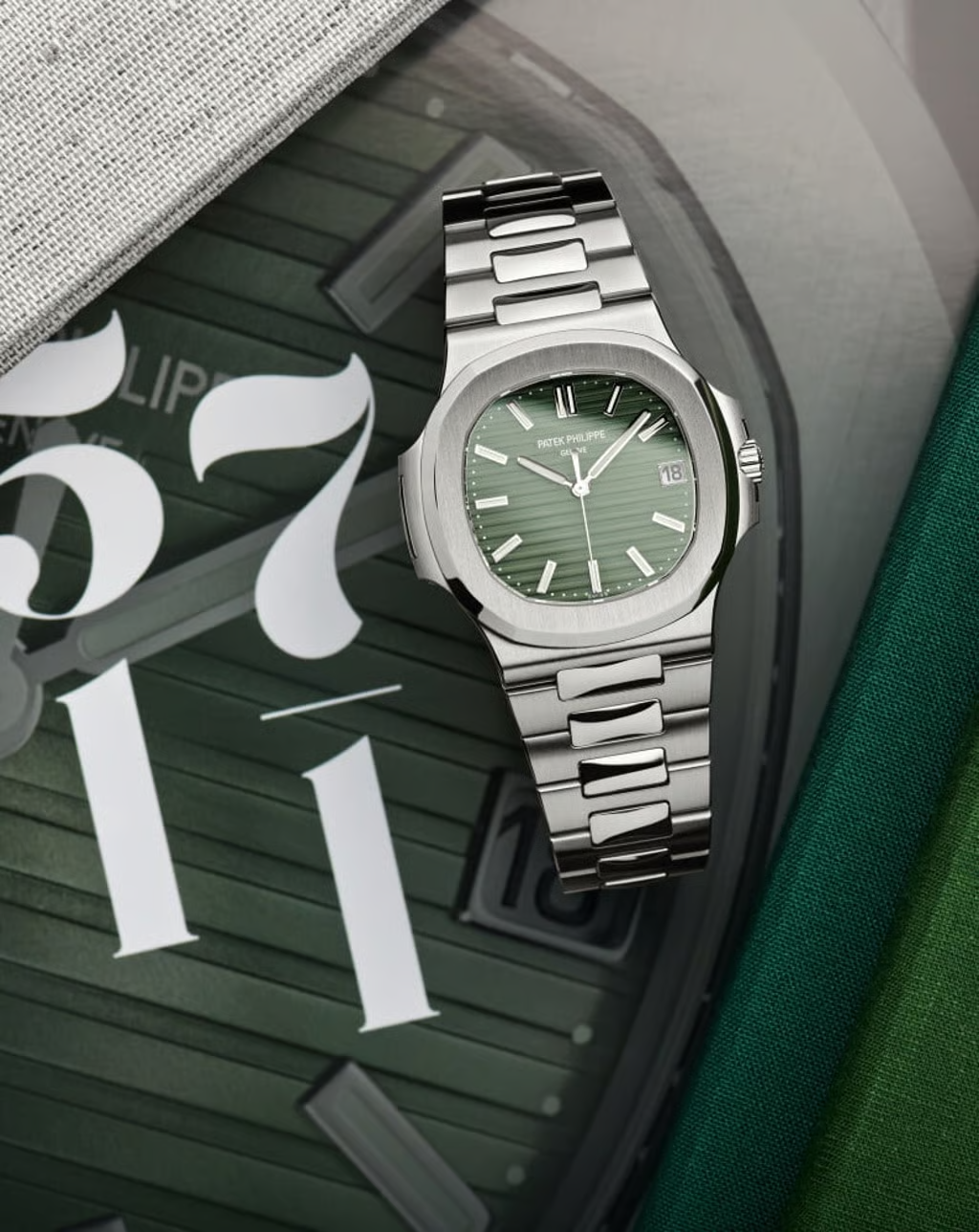 An olive green dial Patek Philippe Nautilus 5711/1A-014 stainless steel luxury watch positioned diagonally over an enlarged graphic print of its dial face and the numbers 5711.