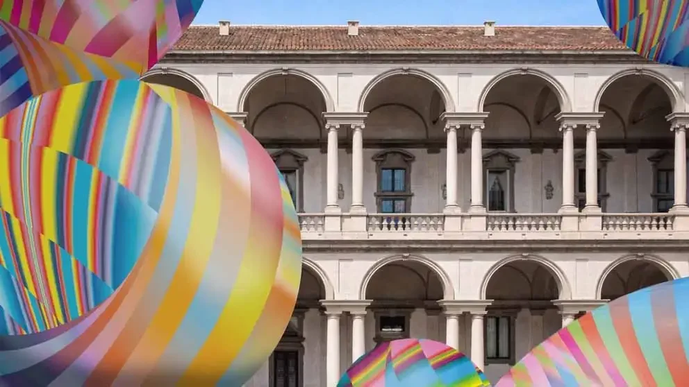 An immersive installation titled Serotonin – the chemistry of happiness by Sara Ricciardi and American Express in the Loggia of the Pinacoteca di Brera, featuring large striped inflatable spheres against classical Italian architecture.