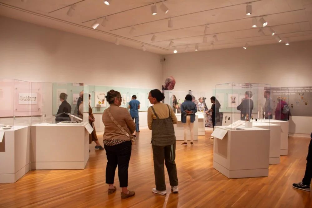 Visitors viewing jewelry and mixed-media artworks displayed in glass cases during the exhibition Objects of Affection: Jewelry by Robert Ebendorf, installed in a museum gallery with wood floors and soft lighting.