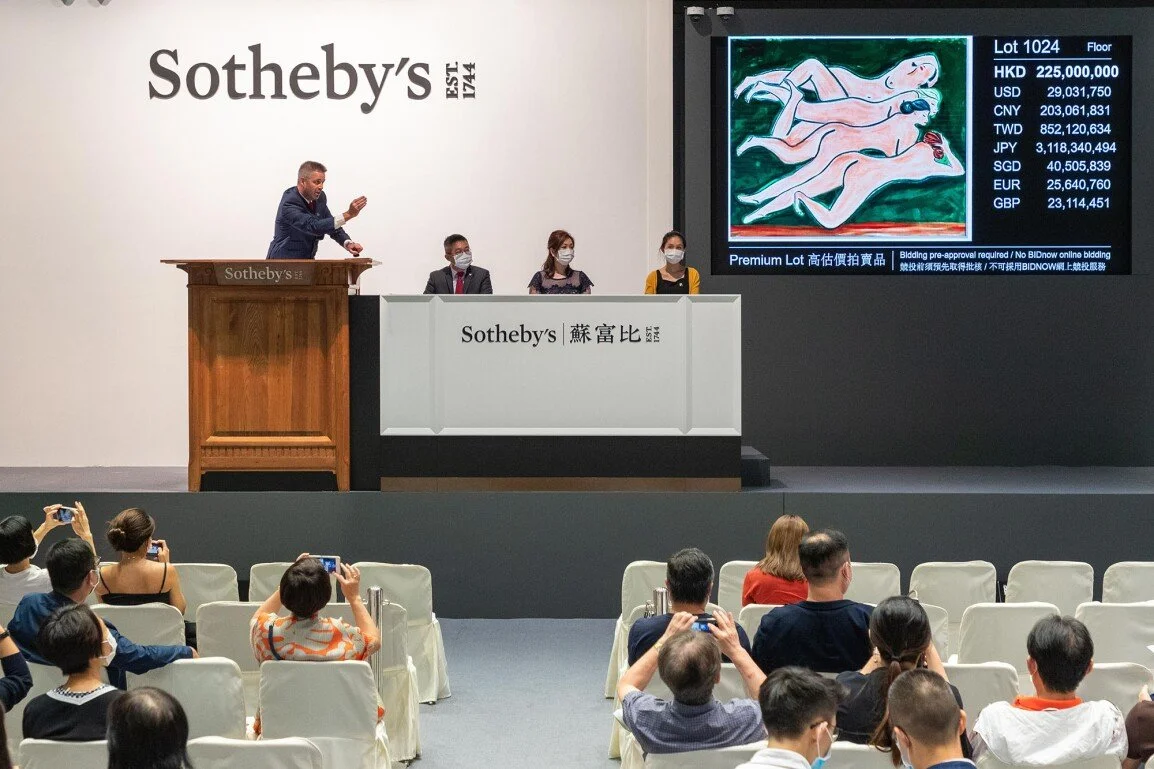 An auctioneer at a Sotheby's art auction, with a painting displayed on a large screen alongside rapidly changing, high bidding prices in multiple currencies, illustrating the speculative nature of the art market.