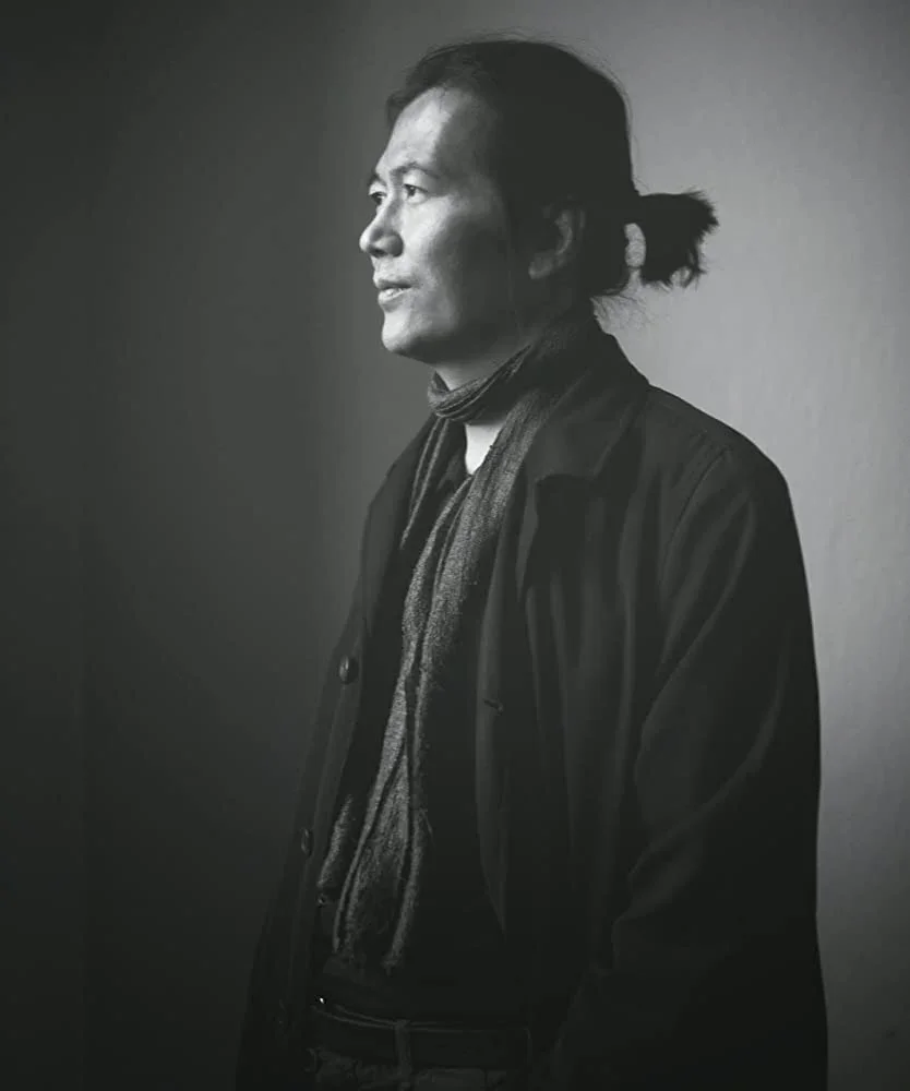 A striking black and white portrait of philosopher Byung-Chul Han, shown in profile with his hair tied back, looking thoughtfully into the distance.