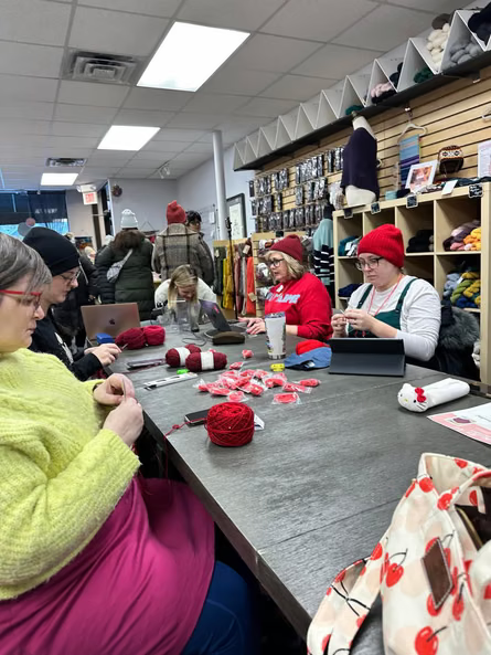 A group of people sitting around a large wooden table inside the Needle and Skein yarn shop in St. Louis Park knitting red Melt the ICE hats surrounded by skeins of red yarn and crafting supplies.