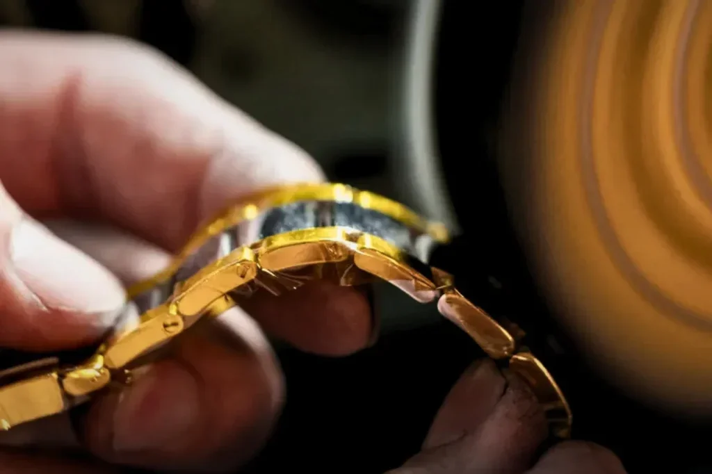 A blurred, high-speed polishing wheel interacts with the gold links of a luxury watch bracelet held by a technician, illustrating the industrial obsession with frictionless, mirror-polished surfaces in traditional luxury.