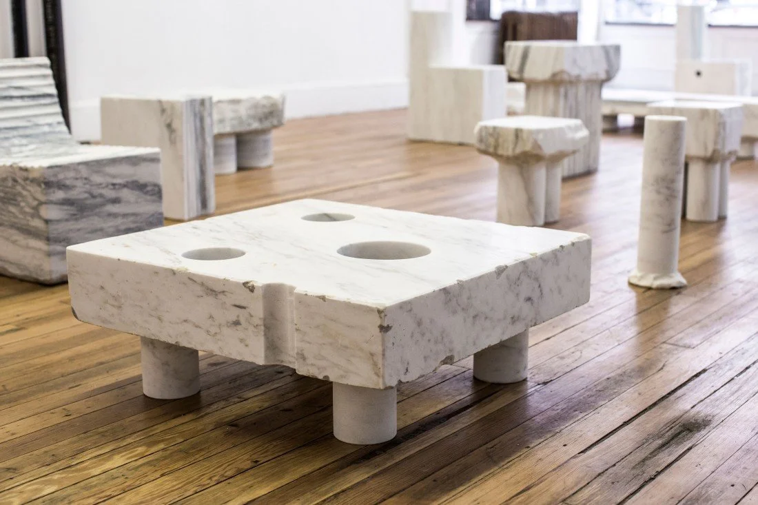 A gallery installation view featuring Max Lamb's monolithic white and gray veined marble furniture, including a low square coffee table with circular cutouts resting on cylindrical stone legs in the foreground.