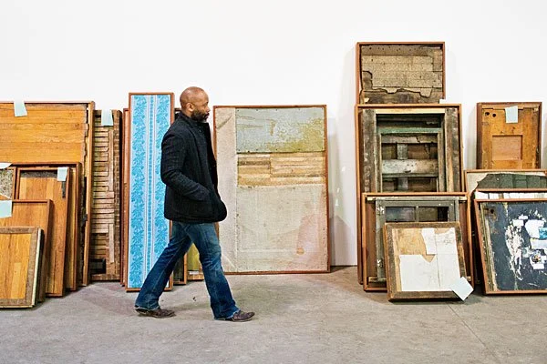 Artist Theaster Gates walking past a series of large-scale archival works and civil tapestries made from reclaimed wood and industrial materials, illustrating the conversion of historical weight into custodial obligation.
