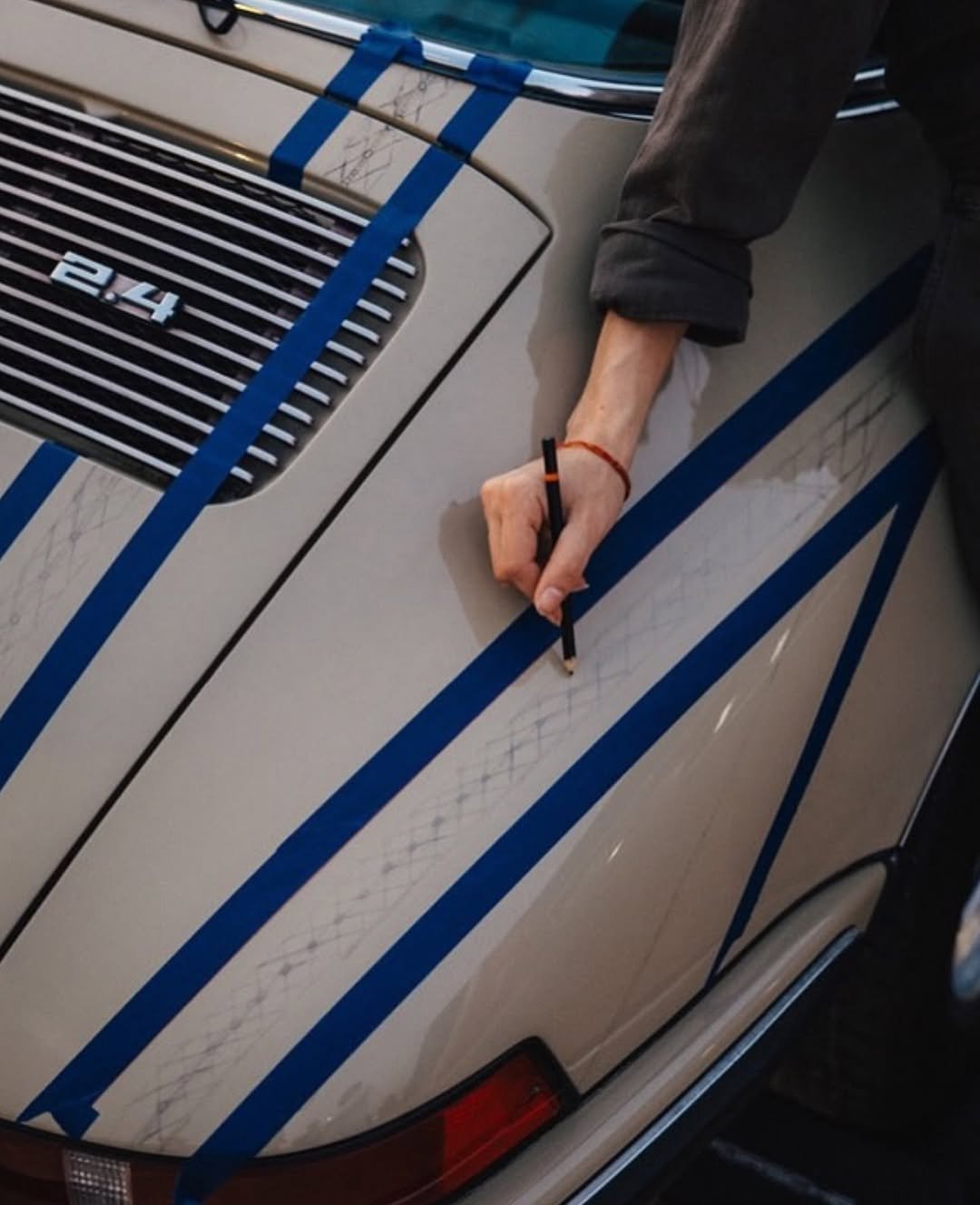 A close-up shot of an artist's hand using a pencil to sketch intricate geometric Al Sadu diamond patterns between strips of blue painter's tape on the engine deck lid of a beige classic Porsche 911 Targa.