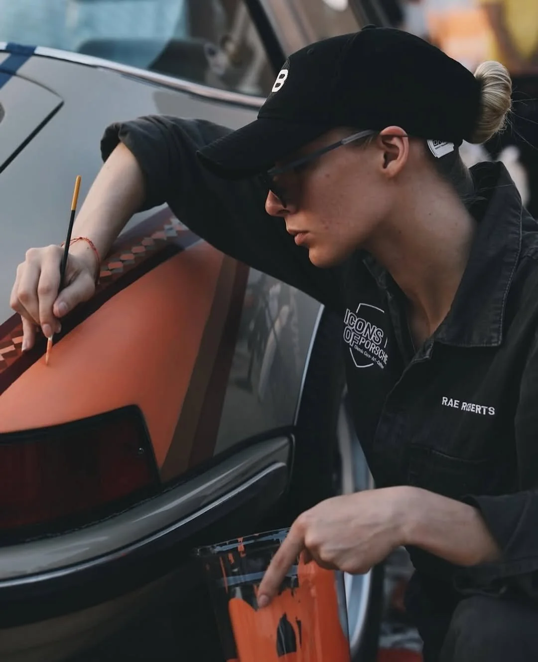 Artist Rae Roberts wearing a black Icons of Porsche work shirt, carefully hand-painting an orange and brown Al Sadu geometric pattern onto the fender of a classic beige Porsche 911 Targa.