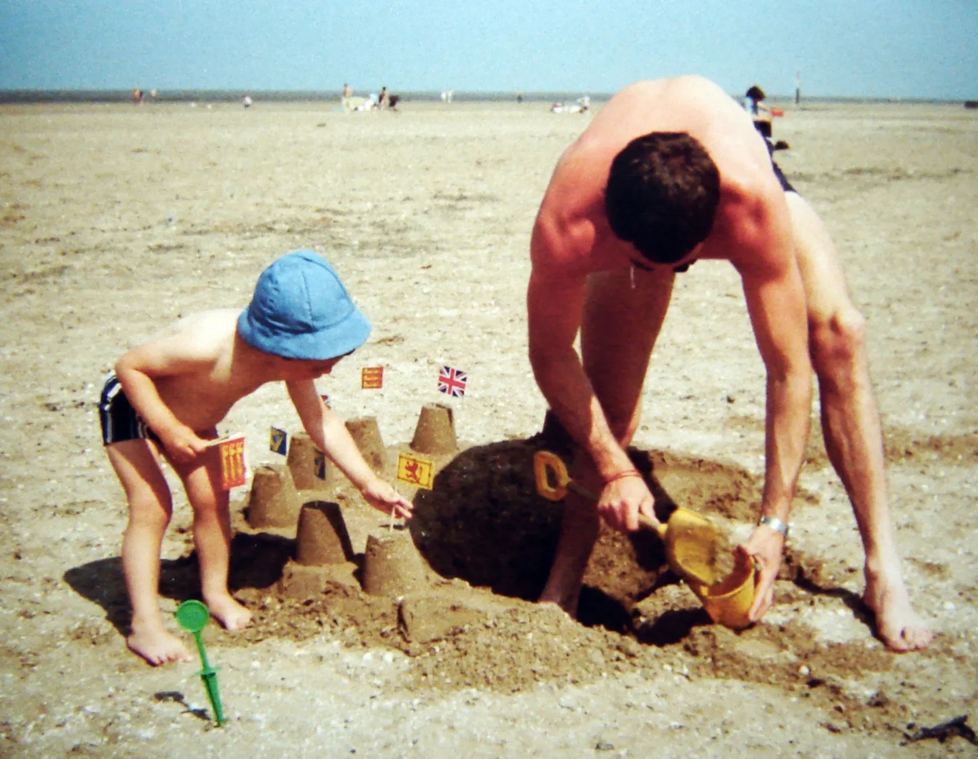A vintage photograph of a young Max Lamb in a blue sun hat building sandcastles with small flags on a wide beach, alongside an adult digging a trench with a plastic shovel.