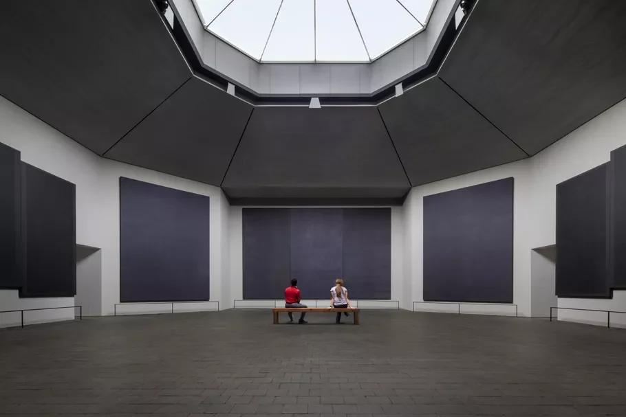 The interior of the Rothko Chapel, showing two people on a bench contemplating the large, dark, immersive canvases in the custom-designed octagonal space, an example of "Mark Rothko's Custodial Strategy."