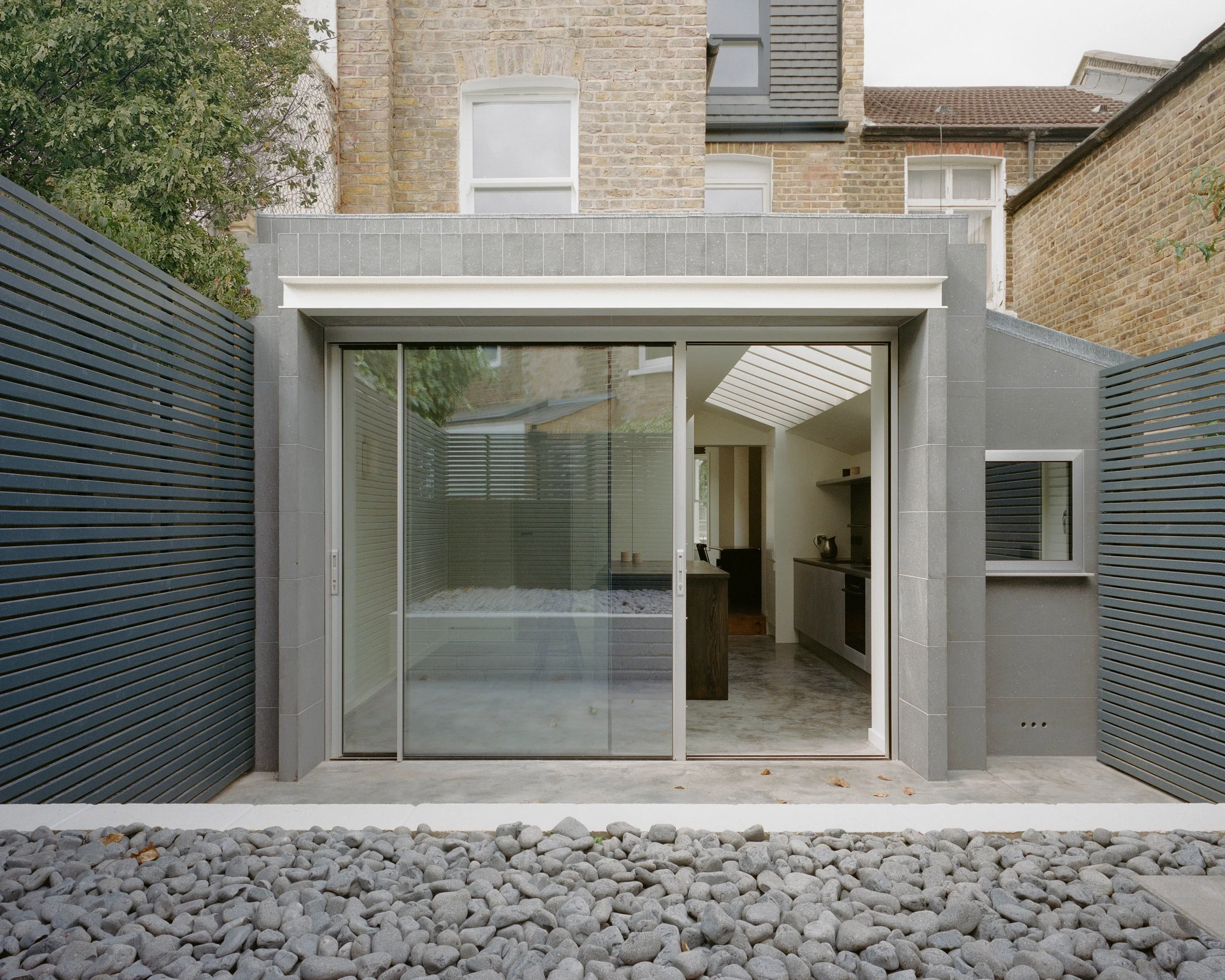 A modern ground-floor extension made of smooth grey basalt stone with large sliding glass doors, juxtaposed against a traditional brick residential building and a pebble-filled foreground.