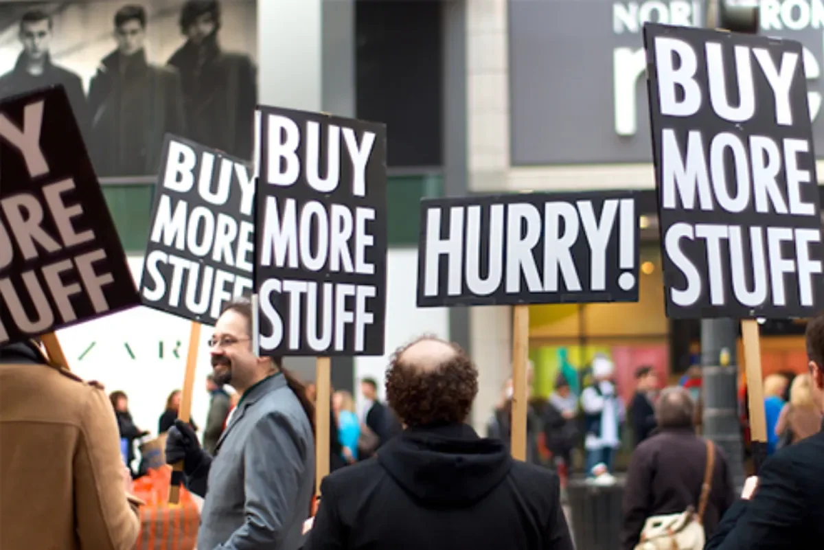 Satirical protest signs reading 'BUY MORE STUFF' and 'HURRY!' held by people on a busy street, symbolizing the ideology of consumer disposability culture.