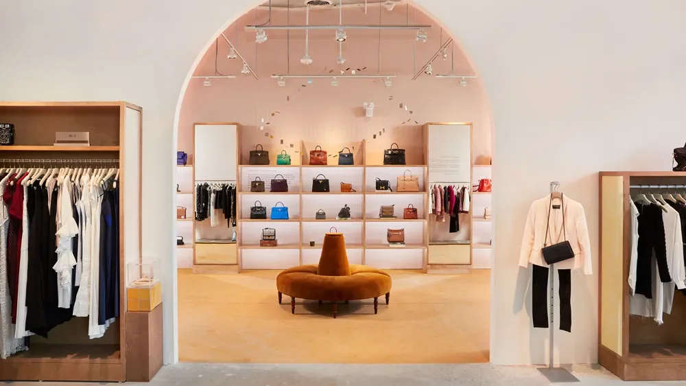 The clean, modern interior of a TheRealReal luxury resale boutique, showing curated displays of high-end clothing and a wall of authenticated Hermès handbags.