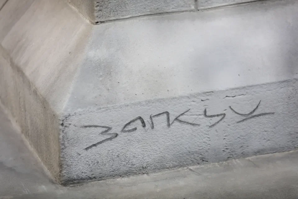 A close up shot of the word BANKSY engraved into the textured gray stone at the base of the Suited Void sculpture plinth in Waterloo Place.