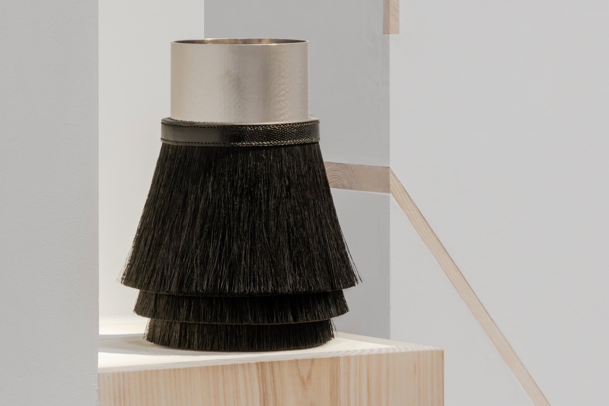 A close-up of a Hermès vessel featuring a hammered palladium neck, a black lizard leather collar, and a tiered horsehair base, situated on a beechwood and plaster plinth.