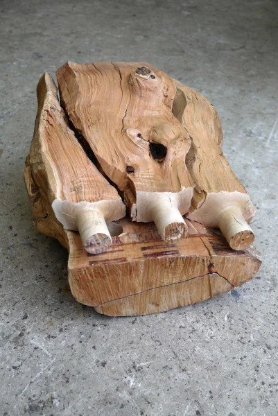 A close-up shot of a Max Lamb raw timber piece resting on a concrete floor, displaying deep structural splits along the wood grain and three hand-carved cylindrical joints protruding outward.