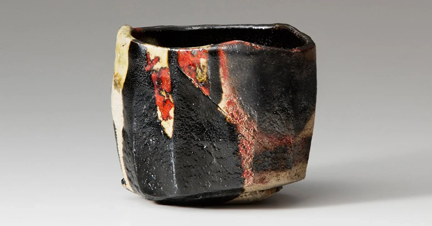 A rustic, irregularly shaped ceramic tea bowl with rough, textured surfaces. It features dark glazes with streaks of red, white, and natural clay, showing visible imperfections and the hand of its maker.