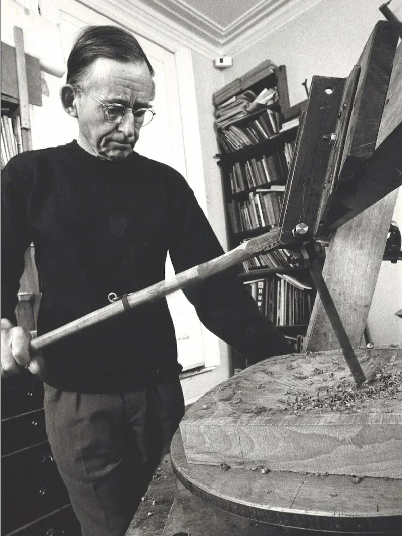 Black and white photo of David Pye in his workshop working on a wooden bowl with hand machinery. David is wearing a black sweater and charcoal pants.