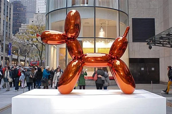 A large, bright orange, highly polished metallic balloon dog sculpture by Jeff Koons stands outdoors on a white pedestal, reflecting its surroundings. Pedestrians walk in the background, some looking at the artwork.