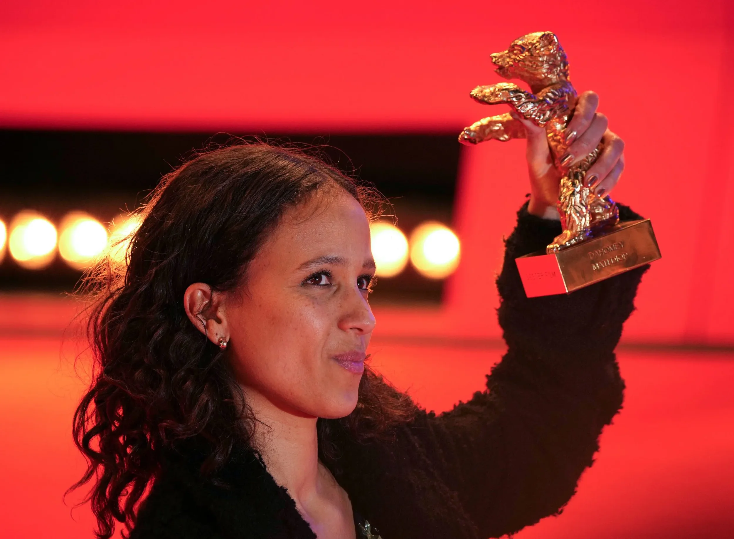 Filmmaker Mati Diop holds up the Golden Bear trophy on stage against a vibrant red background at the Berlin Film Festival for her documentary Dahomey.