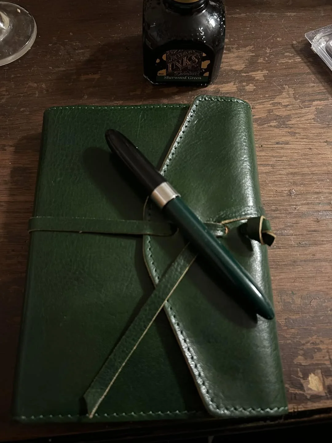 A dark green leather-bound journal with a wrap-around strap and a matching green fountain pen resting on top, set against a rustic wooden surface with a bottle of Sherwood Green ink.
