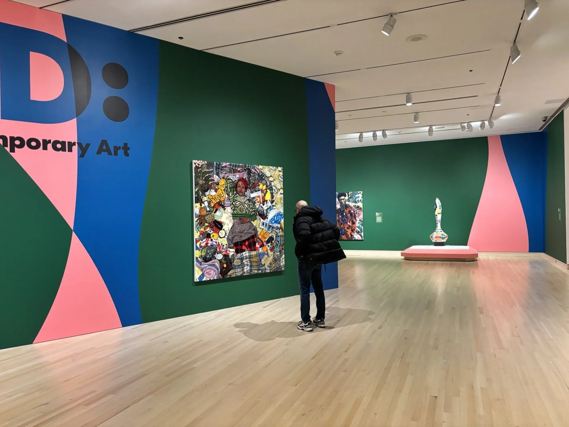 An interior view of the "Bold: New Voices in Contemporary Art" exhibition at the Indianapolis Museum of Art at Newfields, showing a visitor viewing mixed-media contemporary paintings on vibrant green and blue walls.
