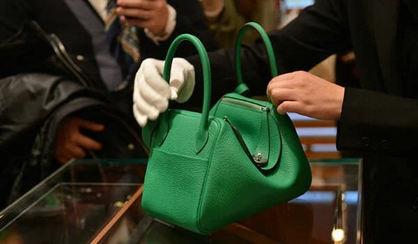 A close-up shot of a person in a black suit and a single white glove meticulously handling a green Hermès Bolide bag on a glass display case to demonstrate luxury custodial care.