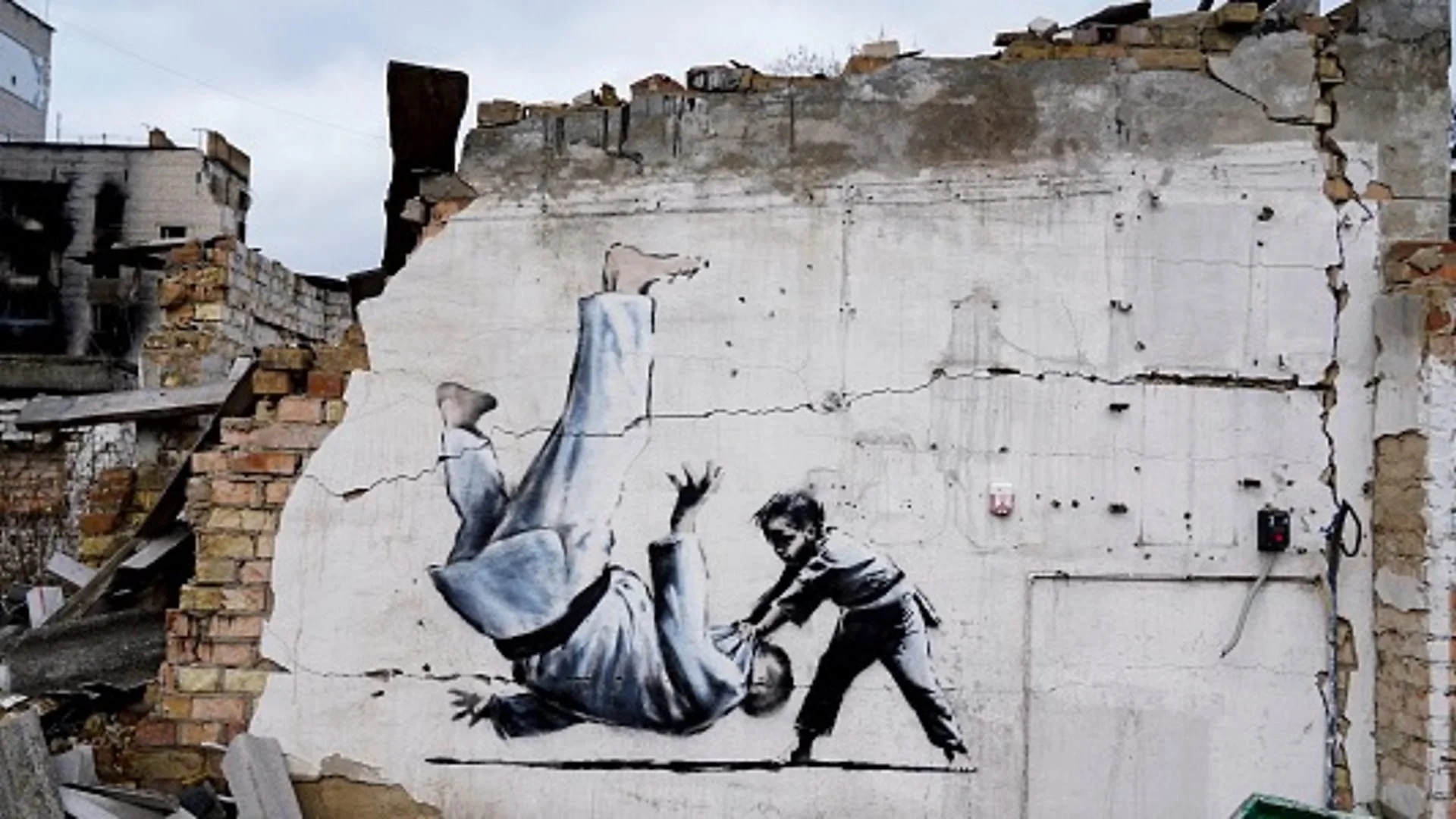 A stencil mural by Banksy on the side of a damaged apartment building in Horenka, Ukraine, depicting a young boy flipping an adult male in a martial arts uniform, symbolizing the David and Goliath nature of the conflict.
