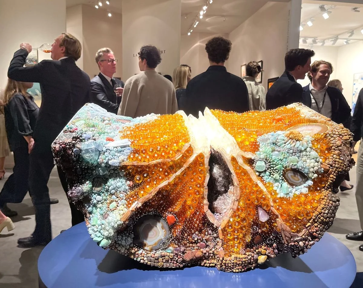 Kathleen Ryan's Bad Fruit gemstone sculpture on display at TEFAF 2026, showcasing dense mineral encrustations simulating an oversized moldering orange slice on a blue pedestal amidst fair attendees.