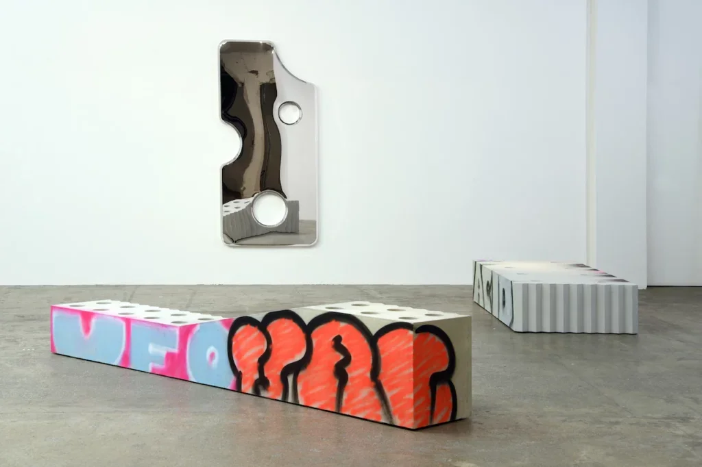 Two low industrial-style sculptural benches with graffiti surfaces and perforated tops, placed before a mirrored wall piece in a minimalist gallery—merging street aesthetics with refined material craft.