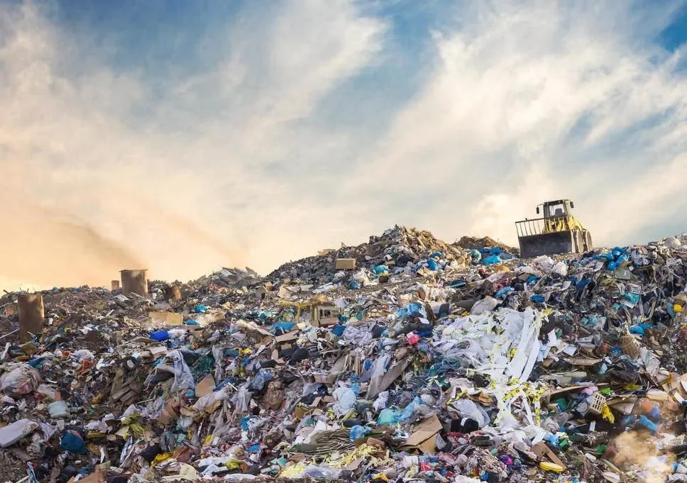 A massive landfill piled high with mountains of garbage and waste under a wide sky. A bulldozer is visible on top of the heap, managing the refuse.
