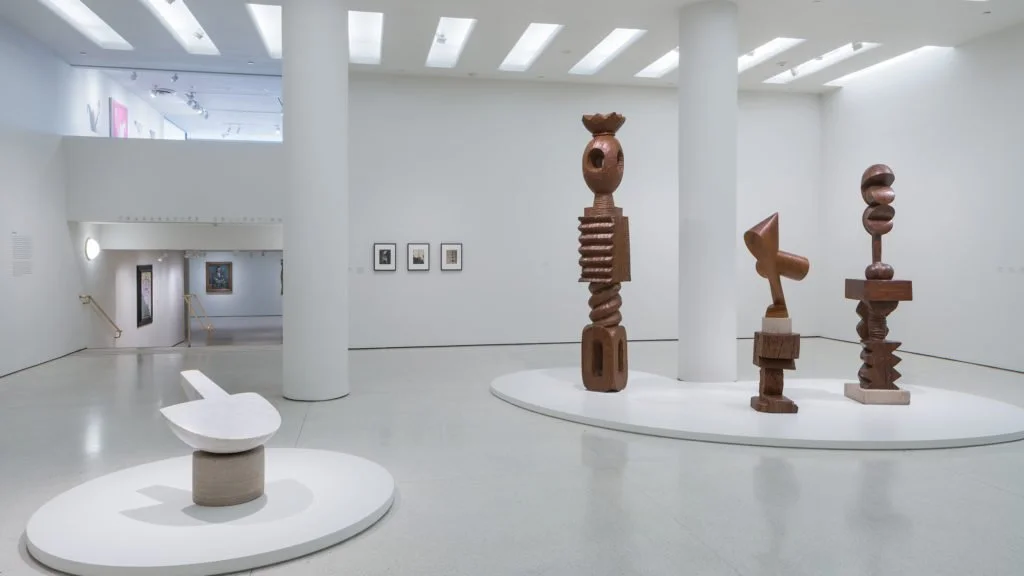 A pristine museum gallery displaying abstract modernist wood and bronze sculptures by Constantin Brancusi on minimalist white pedestals.