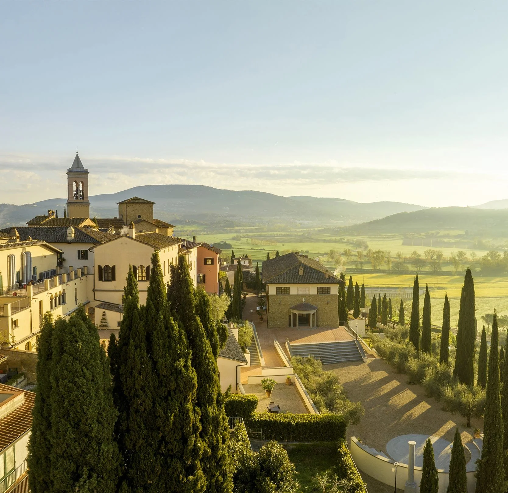 A sunlit wide-angle view of the restored hamlet of Solomeo, Italy, the headquarters of Brunello Cucinelli, showing classic Umbrian architecture, cypress trees, and green valleys beyond.