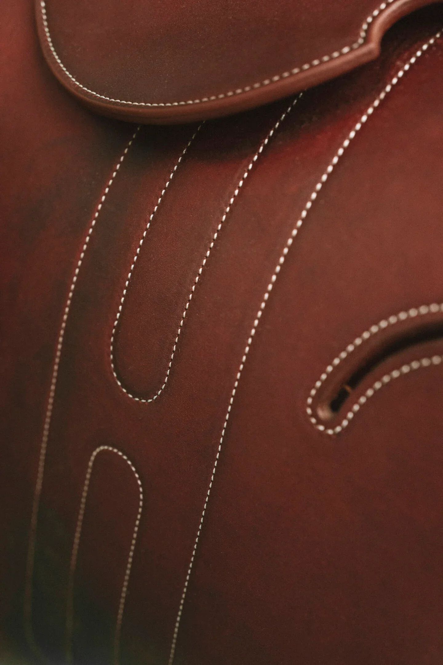 A macro close-up view of a rich brown Hermès leather equestrian saddle, highlighting the precise, parallel lines of signature white hand-executed saddle-stitching.