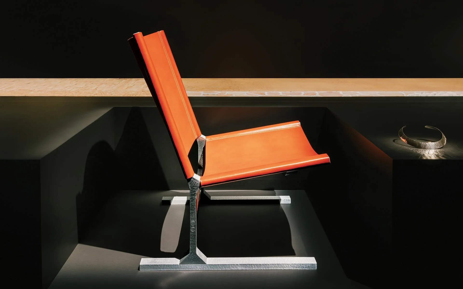 A side profile of a Hermès Palladion chair featuring vivid orange leather and a textured hammered metal frame, photographed by Maxime Verret in a minimalist dark setting at La Pelota during Milan Design Week 2026.