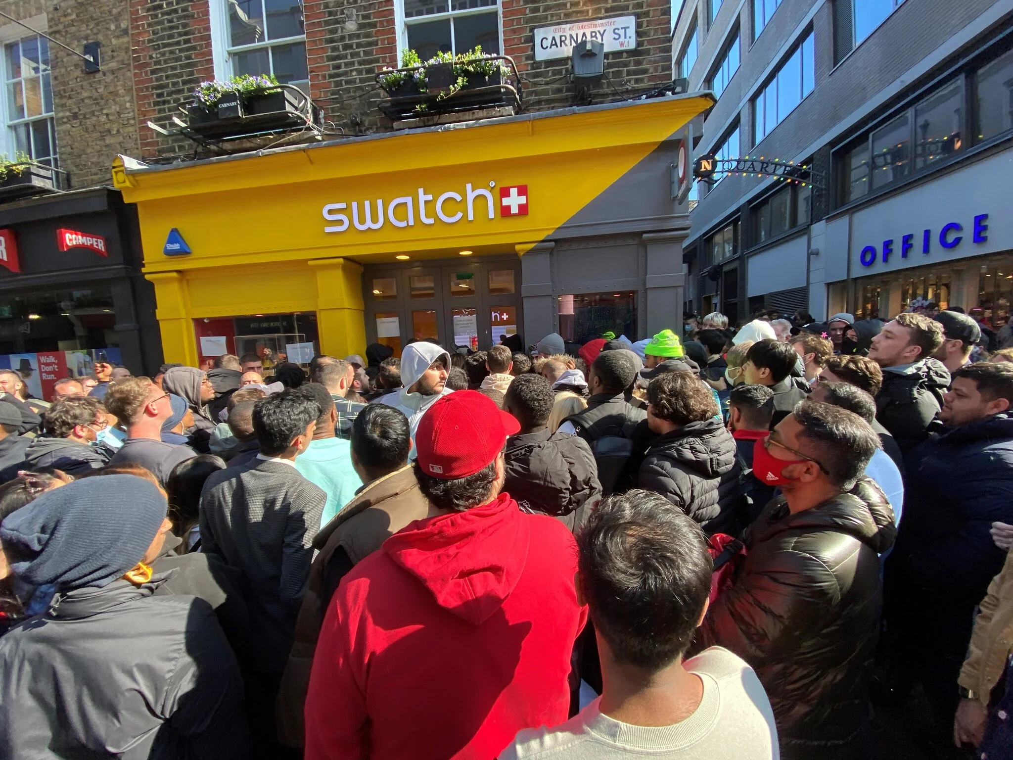 A dense crowd of people gathered outside a yellow and grey Swatch store on Carnaby Street in London during a high-profile watch collaboration launch.
