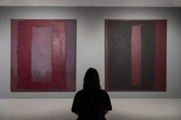 The back of a person with long dark hair viewing two large, dark, color-field paintings by Mark Rothko in a museum setting, demonstrating the contemplative function of the art.