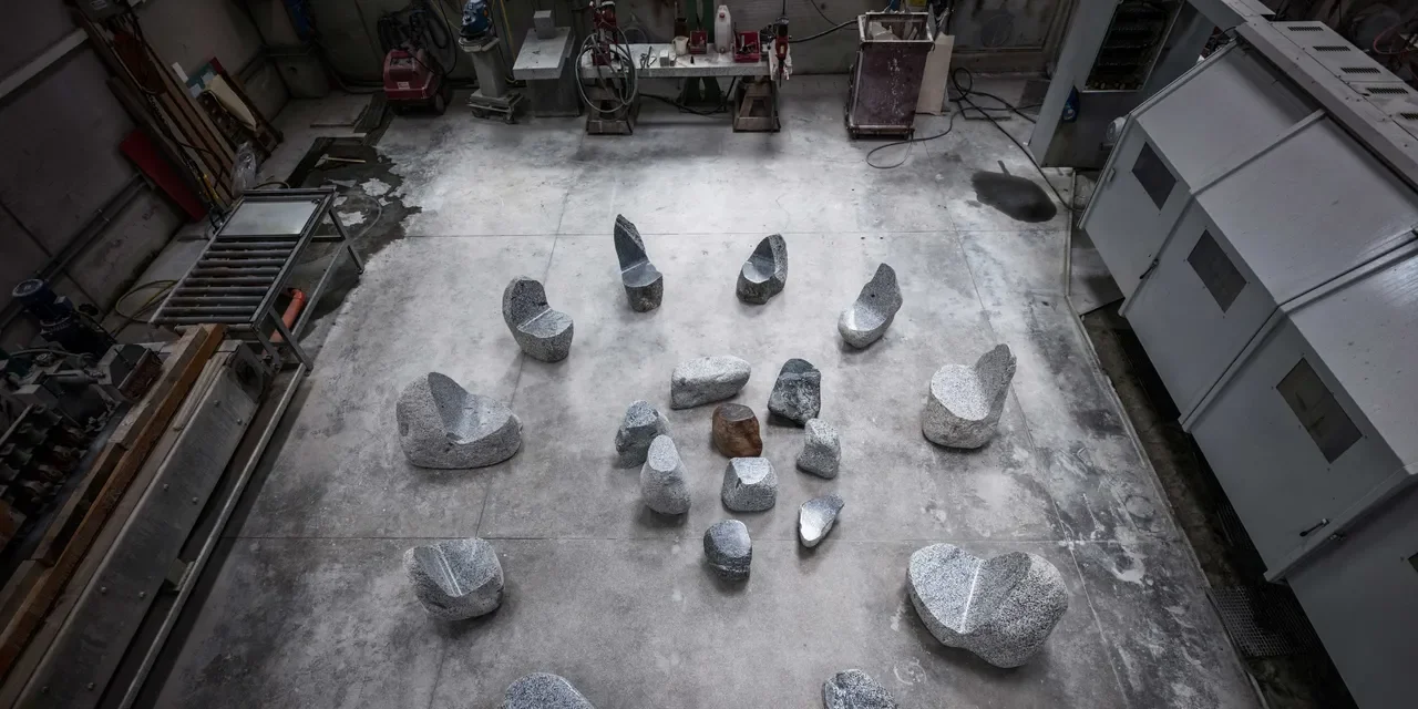 An overhead view of Max Lamb's Elements exhibition pieces arranged in a circular formation on a concrete studio floor, showcasing rough-hewn stone chairs and monolithic rock formations.