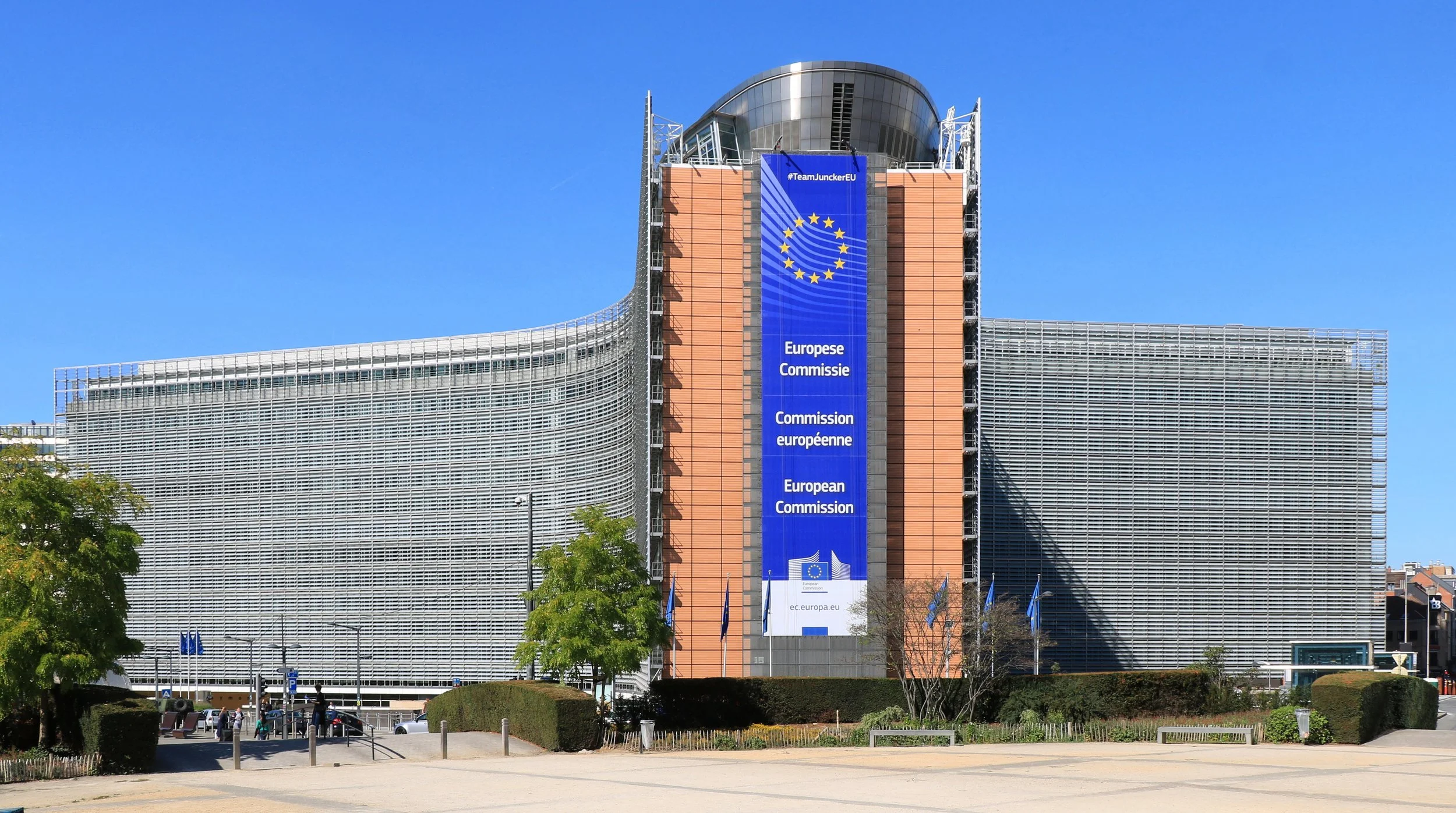 The iconic X-shaped Berlaymont building in Brussels, serving as the headquarters of the European Commission, with a large blue banner displaying the European Union emblem and institutional signage.