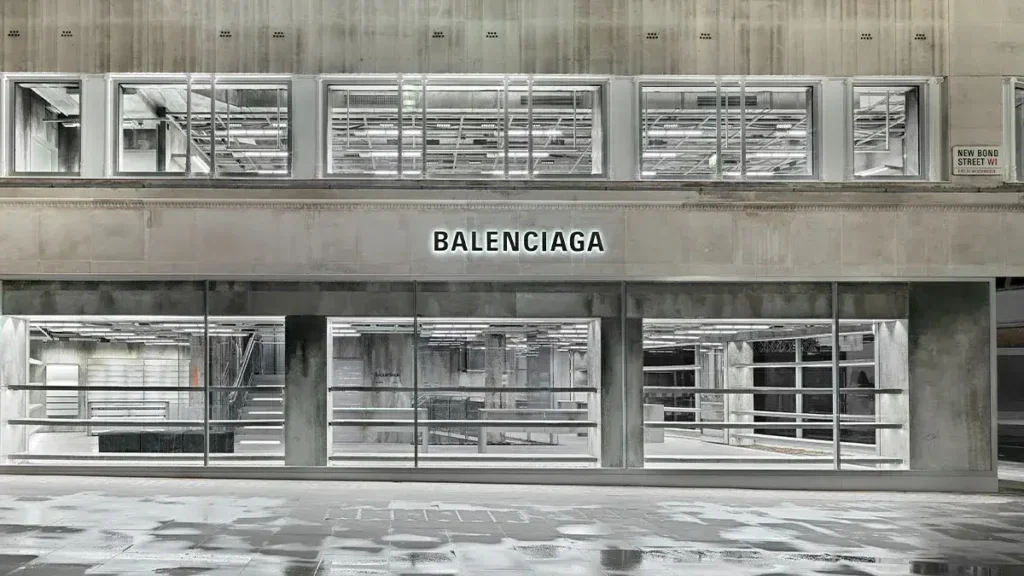 The exterior facade of the Balenciaga flagship boutique on New Bond Street in London, showing a multi-level brutalist design with weathered concrete walls and industrial metal railings under stark lighting.